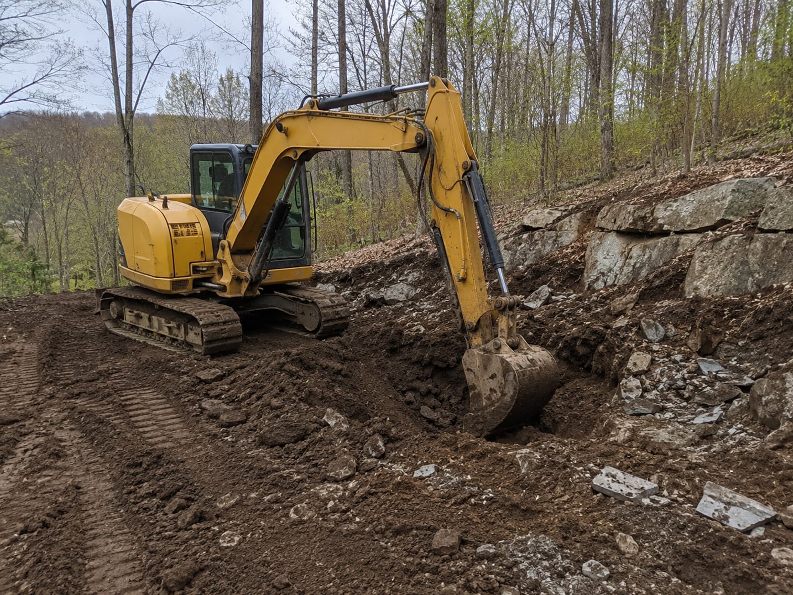 Compact yellow tracked excavator digging into rocky glacial till soil on a sloped residential lot in Watertown CT surrounded by mature deciduous trees