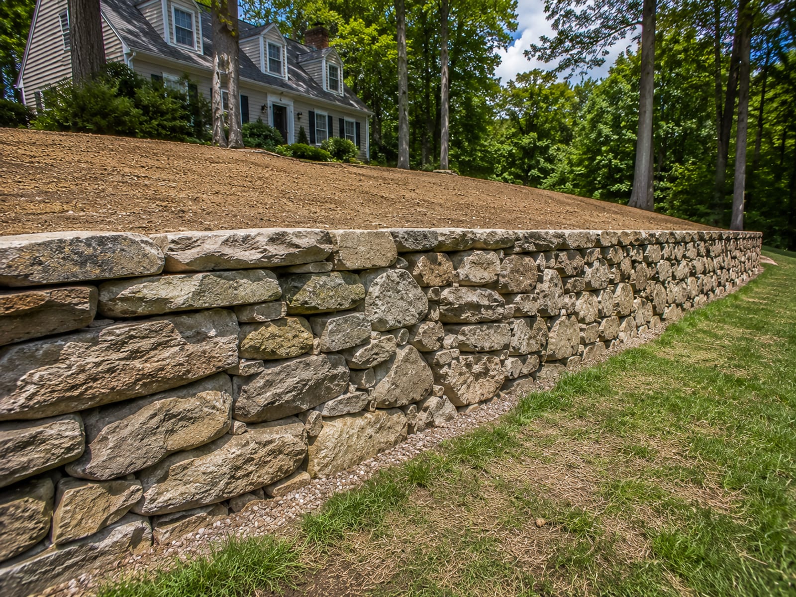 Newly constructed dry-stacked stone retaining wall on a sloped residential property in Wolcott CT with graded earth behind and lawn in foreground