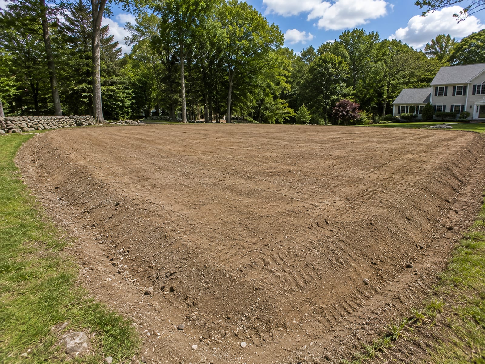 Freshly graded and leveled residential lot in Wolcott CT showing smooth compacted earth ready for construction with colonial home and tree line in background